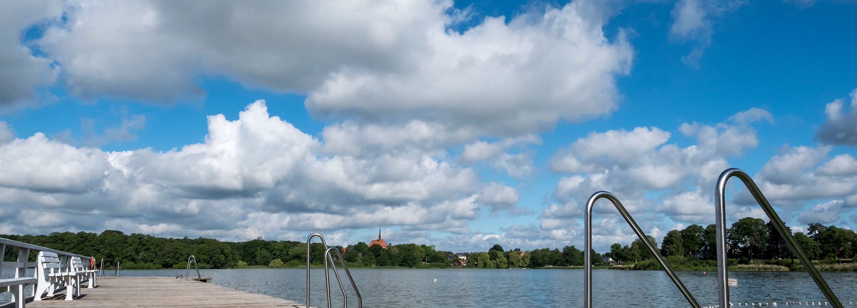Ein Holzsteg führt in den Bordesholmer See im Hintergrund sieht man eine Kirche. Der Himmel ist blau mit Wolken.