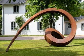 Kupferne Skulptur im Park der NordArt. Im Hintergrund ist ein weißes Gebäude.