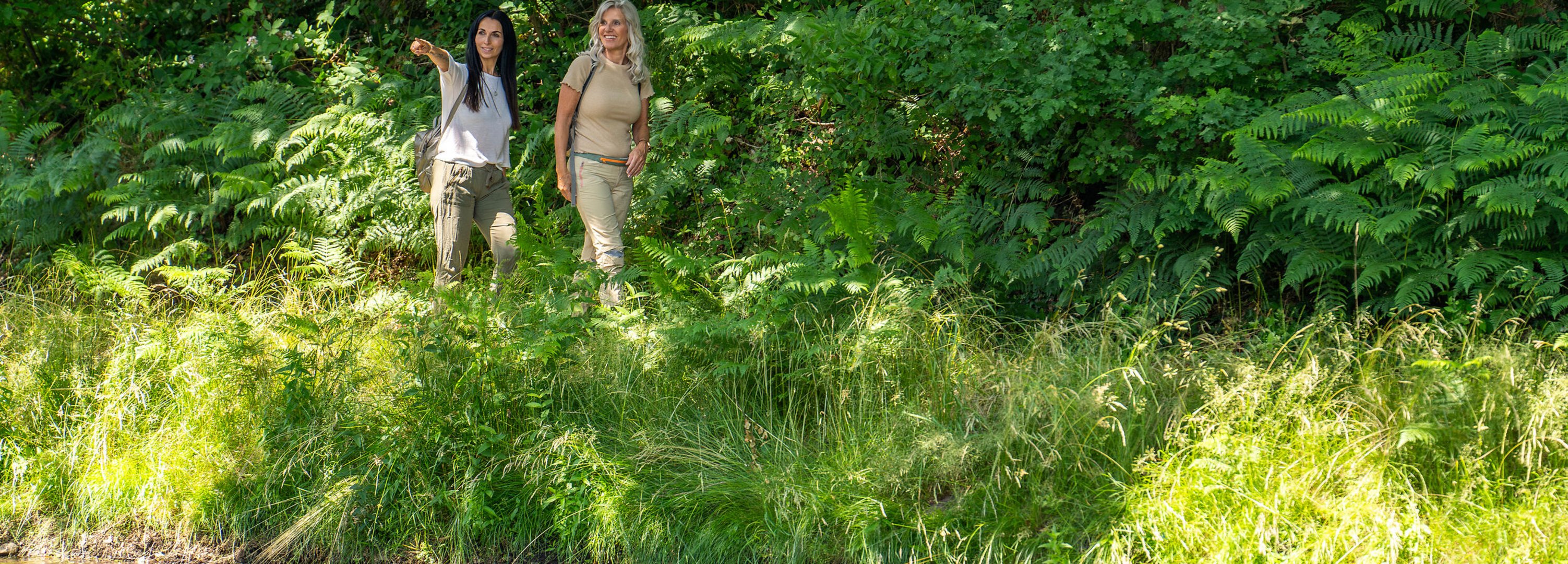 Zwei Frauen wandern durch grüne Landschaft entlang eines Gewässers. 