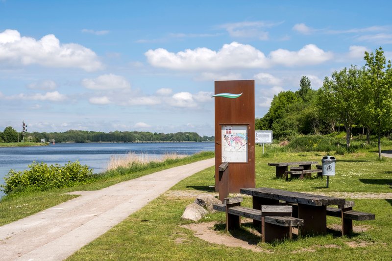 Ein ruhiger Schiffsanleger in Schacht-Audorf mit Picknicktischen und einem Informationsschild am Ufer eines Flusses.
