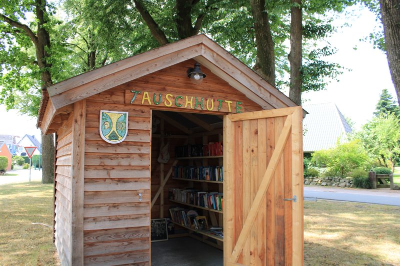 Eine kleine Holzhütte mit der Aufschrift 'Tauschhütte' und einem offenen Eingang, der Bücherregale zeigt.