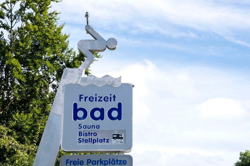 Schild eines Freizeitbads mit Piktogramm eines Tauchers und Informationen zu Sauna, Bistro und Stellplatz.