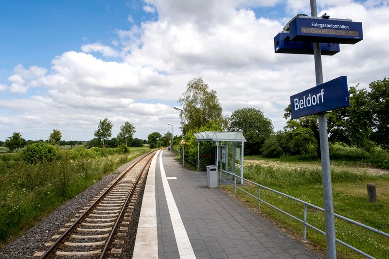 Ein ländlicher Bahnhof mit einem Bahnsteig und Gleisen in Beldorf, umgeben von grüner Landschaft und Bäumen unter einem bewölkten Himmel.