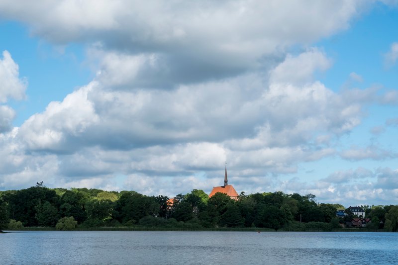 Blick auf den Bordesholmer See mit einer Kirche im Hintergrund und bewölktem Himmel.