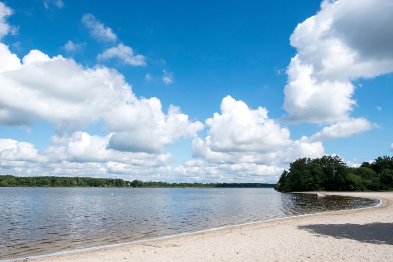 Ein Sandstrand am Einfelder See unter einem blauen Himmel mit weißen Wolken.