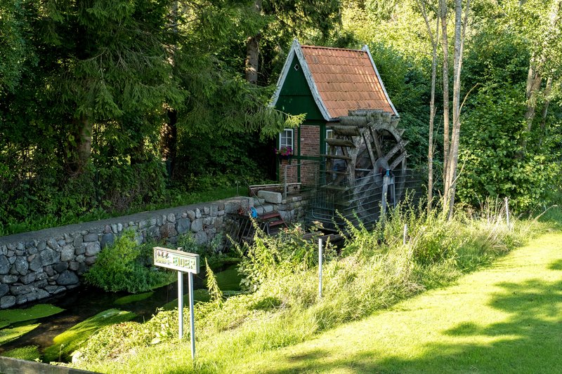 Eine kleine Wassermühle mit rotem Ziegeldach und Wasserrad in einer grünen, bewaldeten Umgebung.