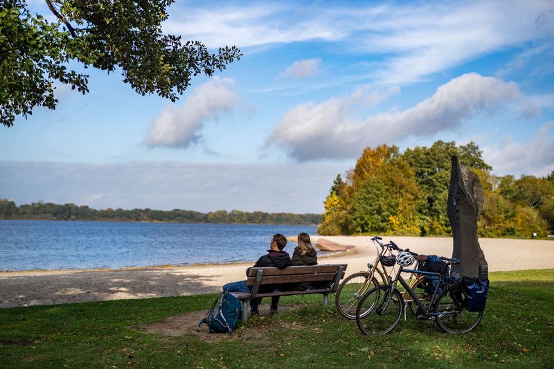 Zwei Personen sitzen auf einer Bank am Ufer des Einfelder Sees, mit Fahrrädern daneben und herbstlichen Bäumen im Hintergrund.
